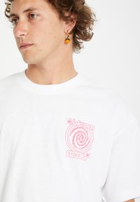 White cotton t-shirt featuring a pink spiral logo with "Volcom" and "STONECO" text. Model has curly hair and an orange beaded earring.