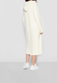 Krämliknande lång hoodie med en fluffig textur och sänkt axel, har en huva och når precis under knäet. Bärs med vita sneakers.