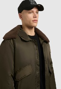 Jacket exterior de culoare verde măsliniu cu guler din blană maro, închidere cu fermoar și buzunare laterale. Purtați cu o șapcă neagră. Textură netedă.