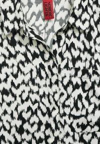 Camicia astratta in bianco e nero con stampa animale, colletto, bottoni frontali, taschino sul petto e etichetta rossa "Nordic Lights" vicino al collo.