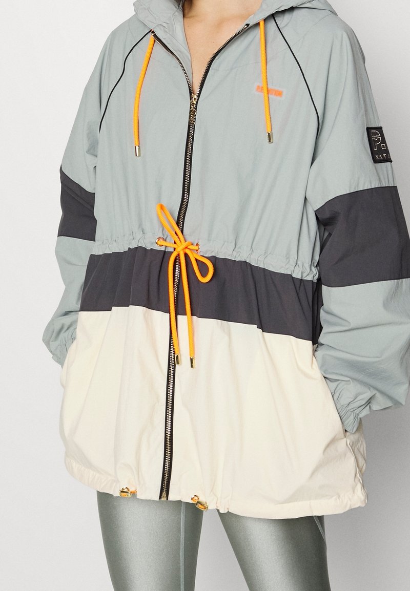 Mujer vestida con una chaqueta cortavientos de bloques de color en gris, gris oscuro y beige, con cordones neón naranja y cremallera plateada, con las manos en los bolsillos.