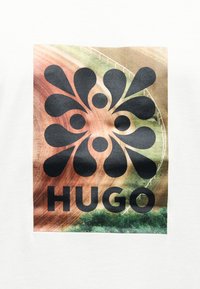 Μαύρο αφηρημένο σχήμα λουλουδιού πάνω από τη λέξη «HUGO» πάνω σε μια υφασμάτινη, πολύχρωμη επιφάνεια με πράσινους και καφέ τόνους.