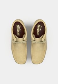 Beige suede shoes with a rounded toe, a tonal lace detail, and a cushioned insole featuring "Clarks ORIGINALS" branding inside.