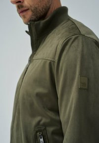 Olivnozelena jakna z gladko teksturo, zip ovratnikom in stranskim žepom. Ima znamčenje na rokavu.