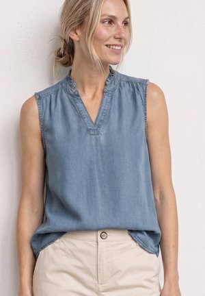 Femme souriante aux cheveux blonds en chignon bas portant un haut bleu sans manches rentré dans un pantalon beige, debout contre un mur blanc.
