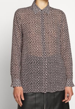 Femme portant une chemise à manches longues boutonnée avec un motif géométrique de triangles noirs et rose clair et une jupe en cuir noire avec fermeture éclair à l'avant.