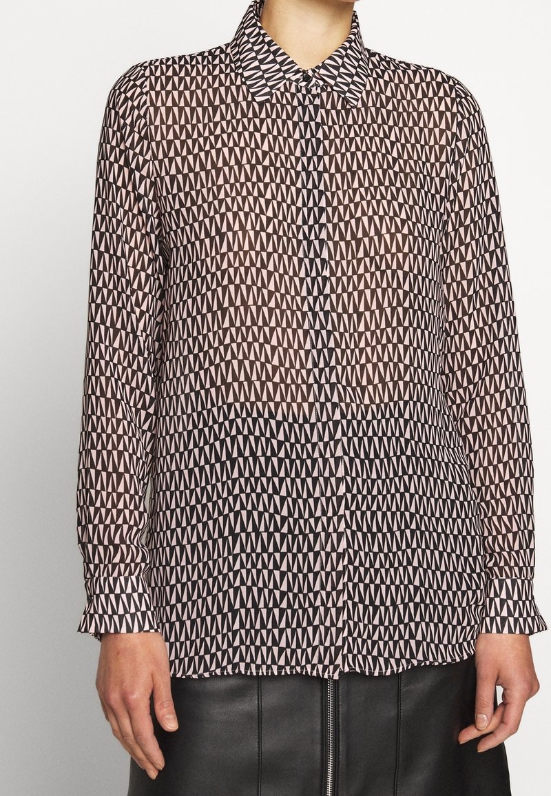 Femme portant une chemise à manches longues boutonnée avec un motif géométrique de triangles noirs et rose clair et une jupe en cuir noire avec fermeture éclair à l'avant.