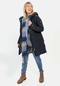 Marineblå pufferjakke med hætte, lynlås og knaplukning, parret med et blå- og hvidstribet tørklæde. Modellen har jeans og brune snørebåndsstøvler på.