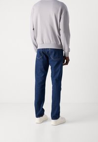 Mand iført en lysegrå sweatshirt, mørkeblå jeans og hvide sneakers, stående med ryggen til en hvid baggrund.