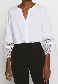 Blusa de algodón blanca con escote en V y detalles de encaje en las mangas acampanadas. Combínala con pantalones negros de corte ajustado para crear contraste.