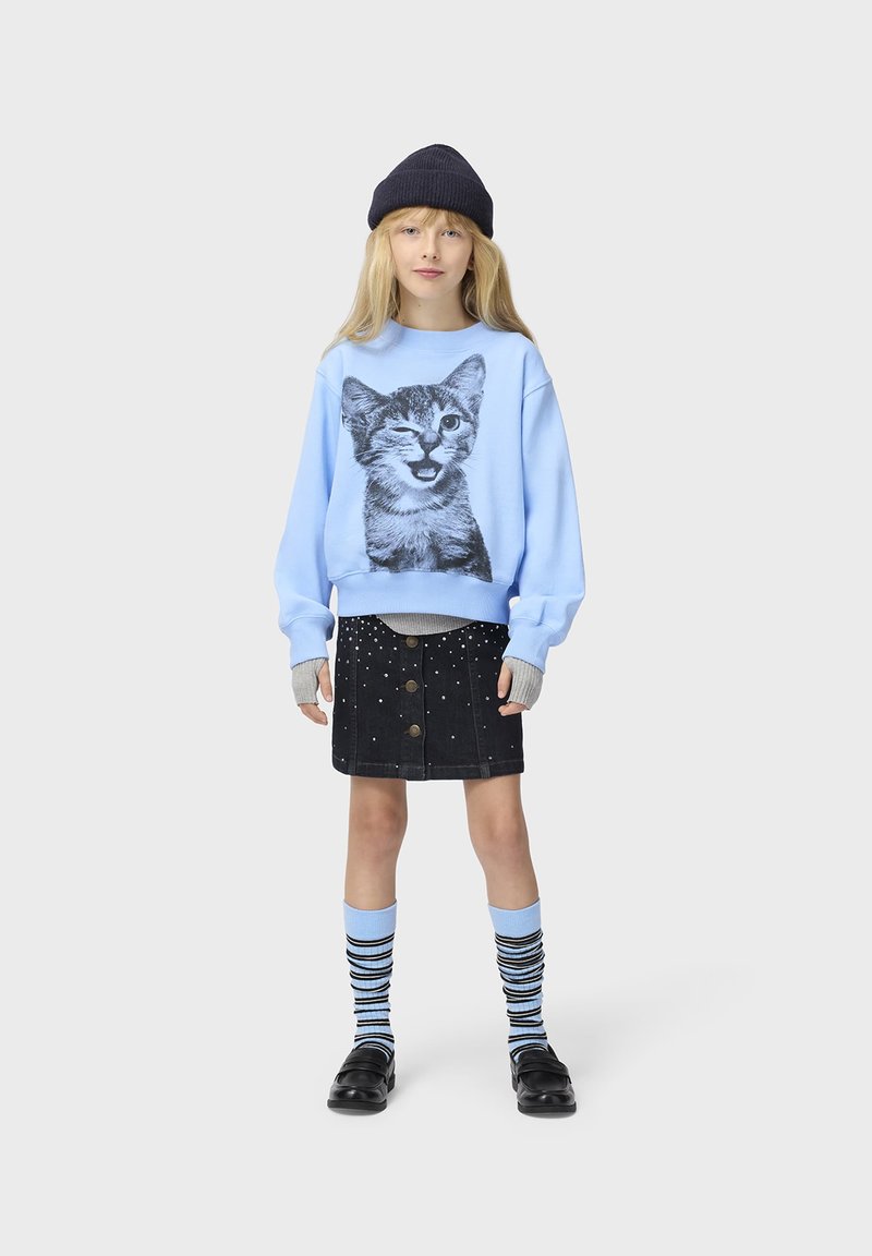 Jeune personne portant un sweat-shirt bleu clair avec un imprimé de chat qui fait un clin d'œil, jupe noire boutonnée avec des perles, chaussettes rayées, chaussures noires et bonnet noir.