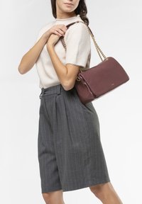 Borsa in bordeaux con texture e tracolla in catena dorata, abbinata a un maglione a maniche corte di un beige chiaro e a pantaloni corti grigi a righe.