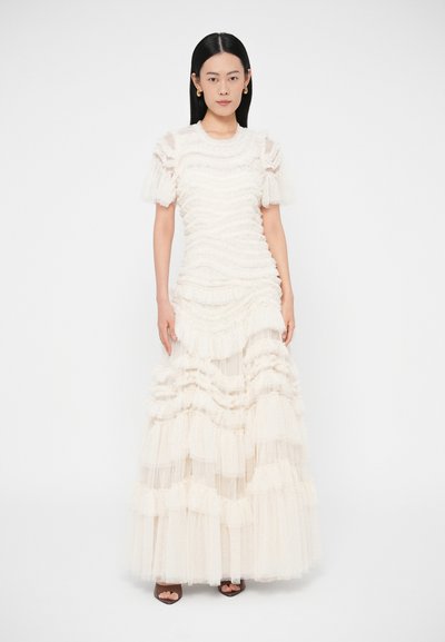 O rochie crem lungă până la podea, cu volane suprapuse, țesătură texturată, mâneci bufante și un corset bine conturat, completată de pantofi cu toc deschiși la vârf.