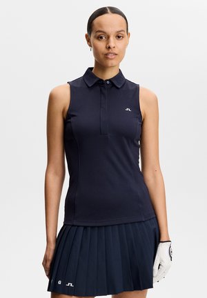 Kobieta ubrana w granatową bezrękawnikową koszulkę polo i plisowaną spódnicę, trzymająca białą rękawiczkę do golfa, na jednolitym tle.