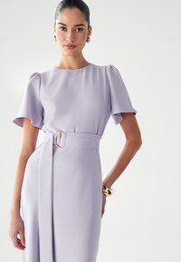 Svetlo lila obleka s kratkimi volanastimi rokavi, oprijetim krojem in pasom z zlatim okroglaškim zapiranjem. Gladka tekstura tkanine.