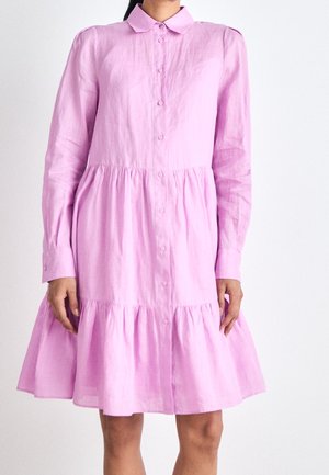 Femme portant une robe rose à manches longues, col, boutonnée, avec une taille froncée et une jupe à volants sur un fond blanc uni.