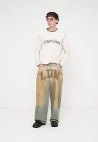 Επιπλέον μεγάλο μπεζ φούτερ με κείμενο «Fatness», συνδυασμένο με πλατιά τζιν παντελόνια σε πράσινο-καφέ απόχρωση με φθαρμένη στάμπα «LDN». Μαύρα χοντρά παπούτσια.