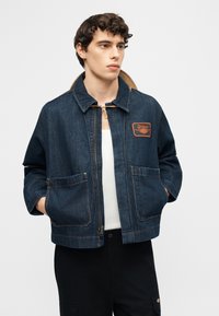 Νέος άνδρας που φοράει σκούρο μπλε τζιν μπουφάν με λογότυπο σε πατς στο στήθος, λευκό πουκάμισο και μαύρο παντελόνι, στέκεται με τα χέρια στις τσέπες του μπουφάν.