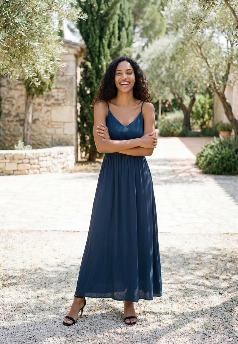 Femme en robe maxi sans manches bleu marine et talons noirs sourit, les bras croisés, en plein air sur un chemin de gravier ensoleillé avec des arbres et des murs en pierre.