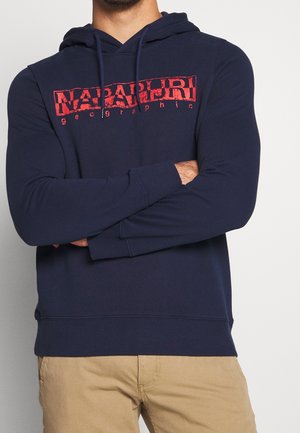 Hombre con sudadera con capucha azul marino con el logo rojo "NAPAPIJRI geographic", con los brazos cruzados, combinado con pantalones beige sobre un fondo liso.
