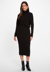 Abito a maglia nero a coste con collo alto, caratterizzato da un design incrociato nella parte superiore. Indossato con stivaletti neri alla caviglia. Tessuto liscio e aderente.