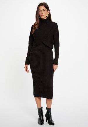 Abito a maglia nero a coste con collo alto, caratterizzato da un design incrociato nella parte superiore. Indossato con stivaletti neri alla caviglia. Tessuto liscio e aderente.