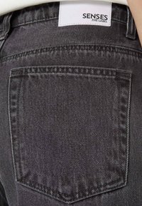 Dark gray denim jeans featuring a back pocket with a subtle textured finish, stitched detailing, and a white label reading "SENSES .THE LABEL."