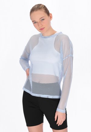 Junge Frau mit zurückgebundenen Haaren, die ein hellblaues, langärmliges Netzoberteil über einem weißen Tanktop und schwarzen Radhosen trägt.