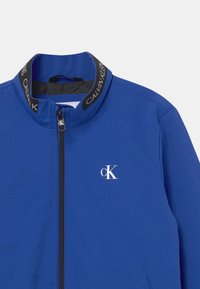 Niebieska kurtka zapinana na zamek błyskawiczny z czarnym kołnierzem, z napisem "Calvin Klein", białym logo "cK" na piersi oraz czarnym uchwytem zamka z inicjałami "cK".