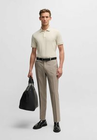 Polo beige, pantaloni marroni chiari, cintura nera, scarpe nere, tiene in mano una grande borsa nera. Design semplice con vestibilità sartoriale.