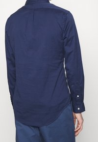 Námořnická košile s dlouhým rukávem a špičatým límcem, zadním skladem a zaobleným lemem. Bavlněný materiál s knoflíkovými manžetami a minimálními texturovými detaily.