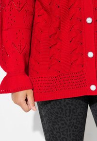 Rote gestrickte Cardigan mit einem strukturierten Muster, gerippten Bündchen und fünf großen silbernen Knöpfen auf der Vorderseite. Getragen über einem schwarzen Leopardenmuster.