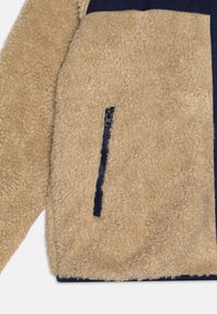 Tan fleecejakke med en marineblå panel øverst. Har en lynlåslomme foran, tekstureret, blødt materiale.