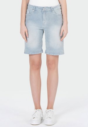 Femme portant un short en jean à rayures bleu clair jusqu'aux genoux et des baskets blanches, debout devant un fond uni.