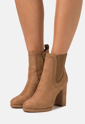 Tan suede ankle boots with a smooth texture, rectangular heel, elastic side panels, and a zipper closure. Features rounded toe design.