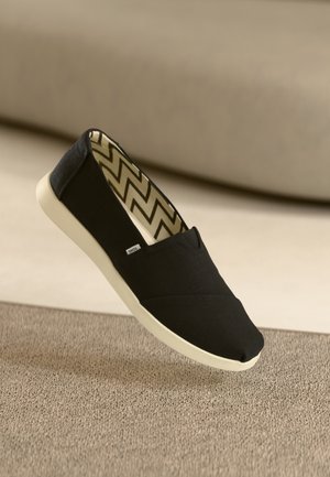 Chaussure noire à enfiler avec semelle blanche et doublure intérieure à motif chevrons posée sur un tapis beige.