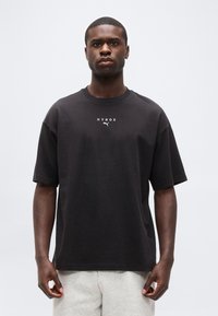 Άνδρας που στέκεται με το πρόσωπο προς τα εμπρός, φοράει μαύρο t-shirt HYROX Puma και ανοιχτό γκρι σορτς σε απλό λευκό φόντο.