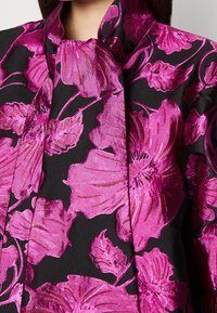 Närbild av svart tyg med stort, glänsande magentafärgat blommönster och hög krage på ett plagg, som visar textur och intrikat broderi.