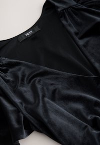 Top in velluto nero dalla texture liscia, con maniche corte a sbuffo e scollo a V. Etichetta "NEXT" visibile vicino al collo.