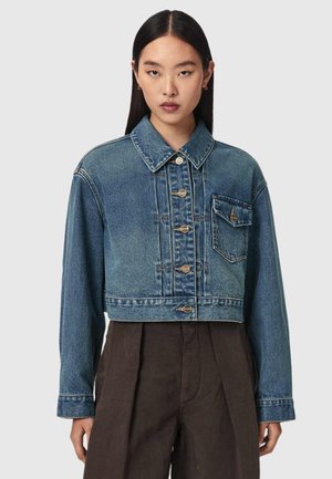 Jonge vrouw met stijl zwart haar draagt een korte blauwe spijkerjas met knopen en donkere broek met hoge taille tegen een effen achtergrond.