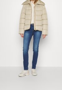 Beige puffer jacket with a large folded collar, zip closure, and horizontal stitching. Worn over light-colored top and blue slim jeans.