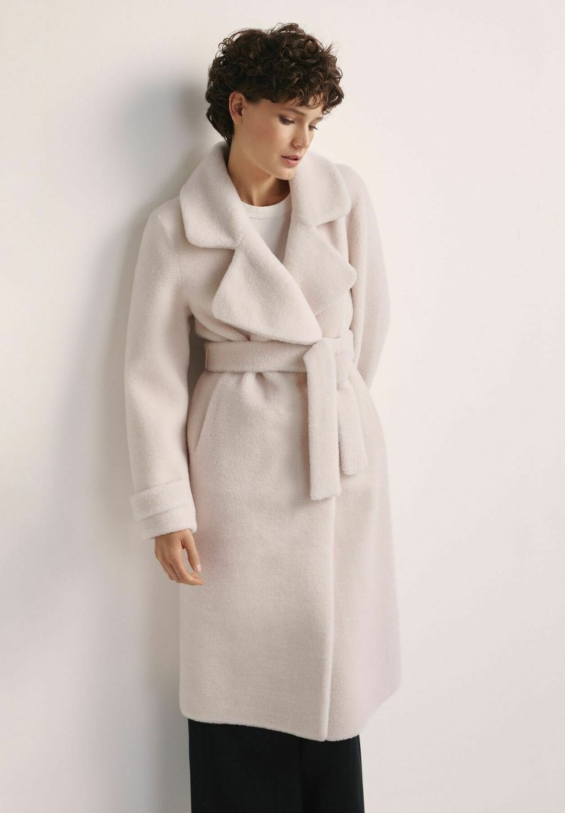 Kobieta z krótkimi kręconymi włosami, ubrana w beżowy płaszcz wełniany z paskiem, na białej bluzce i czarnych spodniach, stojąca na tle gładkiej ściany.