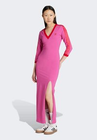 Abito midi aderente rosa con scollatura a V, maniche a tre quarti a righe rosse, spacco laterale e logo ricamato sul petto. Abbinato a sneaker casual.