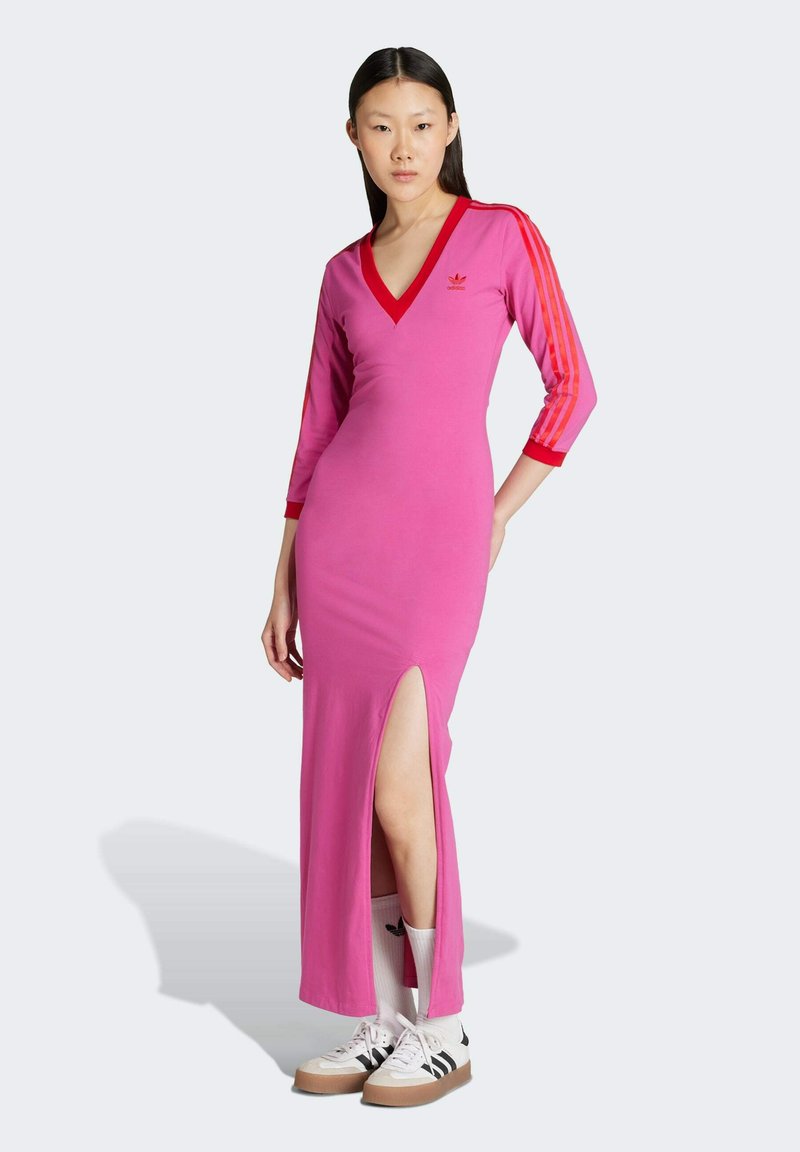 Abito midi aderente rosa con scollatura a V, maniche a tre quarti a righe rosse, spacco laterale e logo ricamato sul petto. Abbinato a sneaker casual.