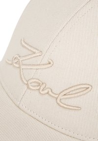 Căciulă de baseball bej, dintr-un material texturat, având un logo "Karl" brodat, reliefat, de o nuanță similară, evidențiat prin detalii de cusătură.