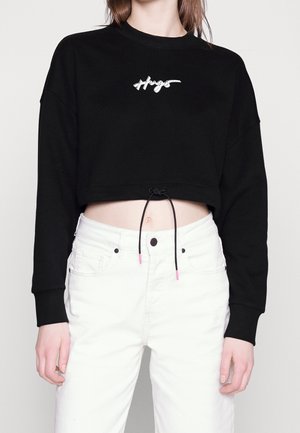 Personne portant un sweat court noir avec le logo "Hugs" et un pantalon blanc taille haute, montrant le ventre et un détail avec cordon.