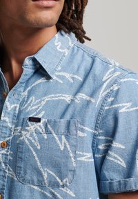 Kurzärmeliges Jeanshemd in Hellblau mit weißem abstraktem Muster. Verfügt über eine durchgehende Knopfleiste, einen Kragen und eine Brusttasche mit Logo.