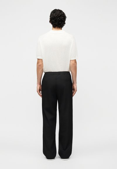 Άνδρας με κοντά σγουρά σκουρόχρωμα μαλλιά στέκεται στραμμένος προς τα πίσω φορώντας λευκό κοντομάνικο πουκάμισο και μαύρο παντελόνι με φαρδιά μπατζάκια και ελαστική μέση.