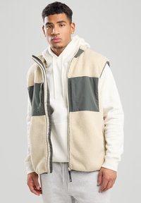 Beige Fleeceweste mit olivgrüner Einsätzen, Reißverschluss, hohem Kragen und Reißverschlusstaschen über einem weißen Hoodie und grauen Jogginghosen.