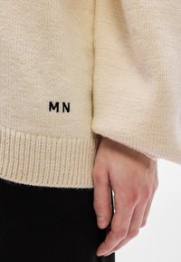 Bézs kötött pulóver bordázott mandzsettákkal és szegéllyel. A szegély közelében fekete "MN" monogram hímzett. Az anyag puha és texturált hatású.